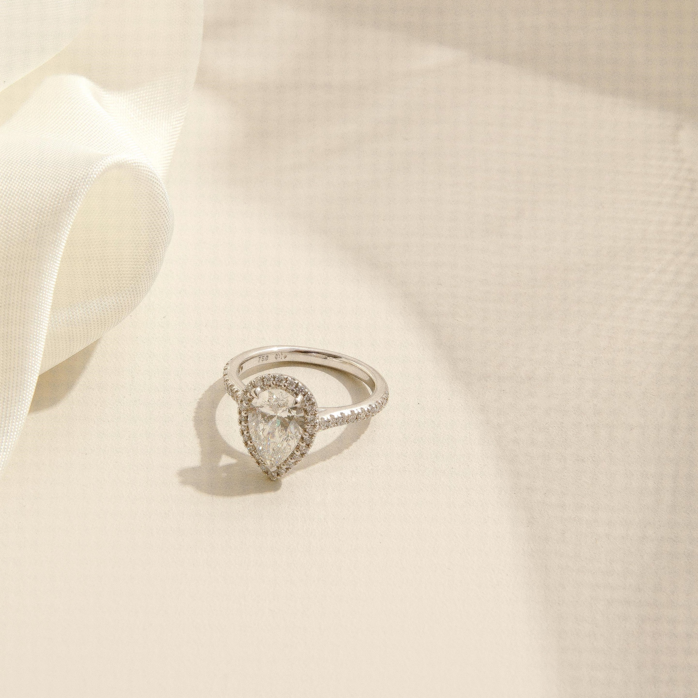 A white gold ring featuring a pear cut diamond surrounded by a halo and pavé band, displayed on a textured cream fabric background with a soft white ribbon. Brisbane Engagement Rings
