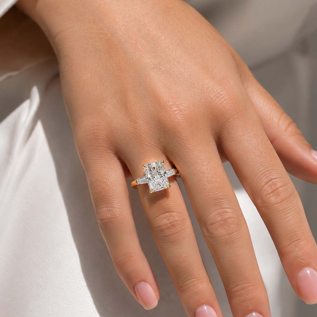 A close up of a hand wearing a yellow gold radiant cut diamond engagement ring and tapered baguette diamonds, photographed against soft white fabric.