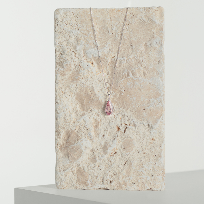 A pink teardrop shaped, diamond encrusted necklace is draped across a stone