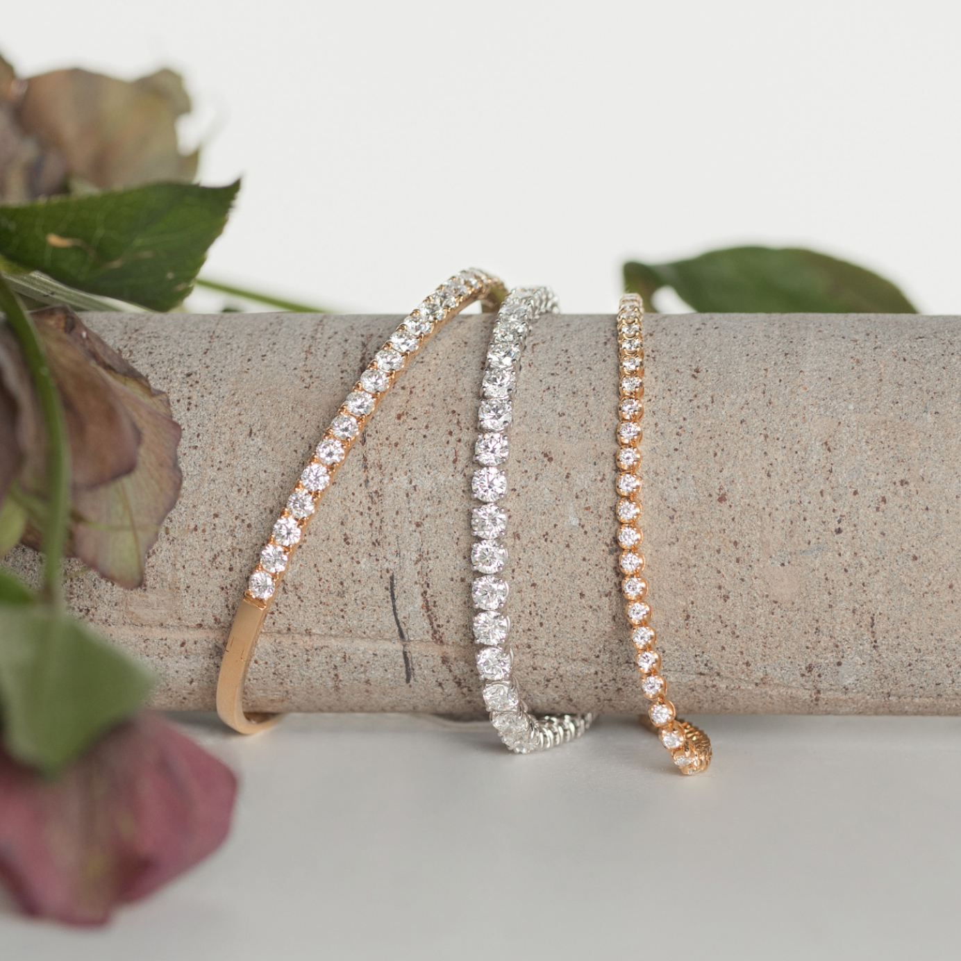 A gold diamond tennis bracelet, gold diamond bangle and a white gold diamond tennis bracelet are stacked on a natural stone column draped with greenery