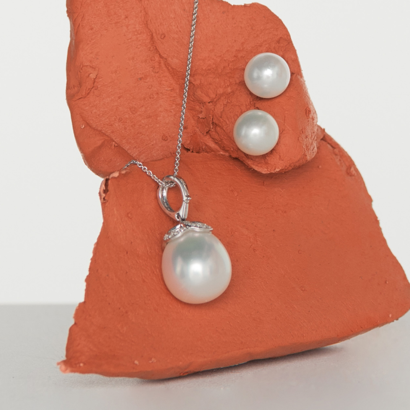 A pearl and diamond necklace and pearl studs are displayed on a piece of terracotta clay